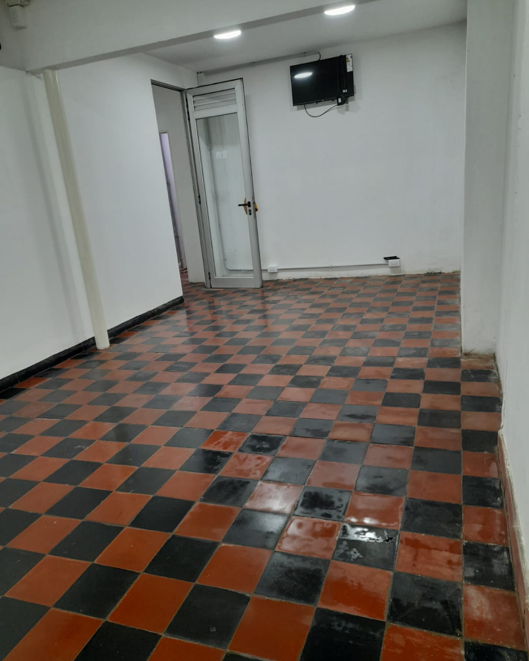 Habitación vacía con suelo de baldosas a cuadros rojos y negros, paredes blancas y un televisor colgado.