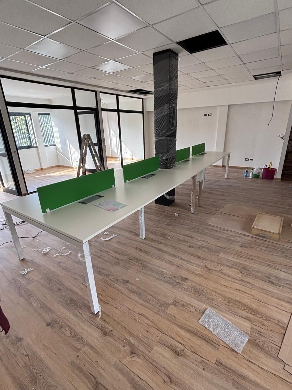 Espacio de oficina con largos escritorios blancos, divisores verdes y pisos de madera.