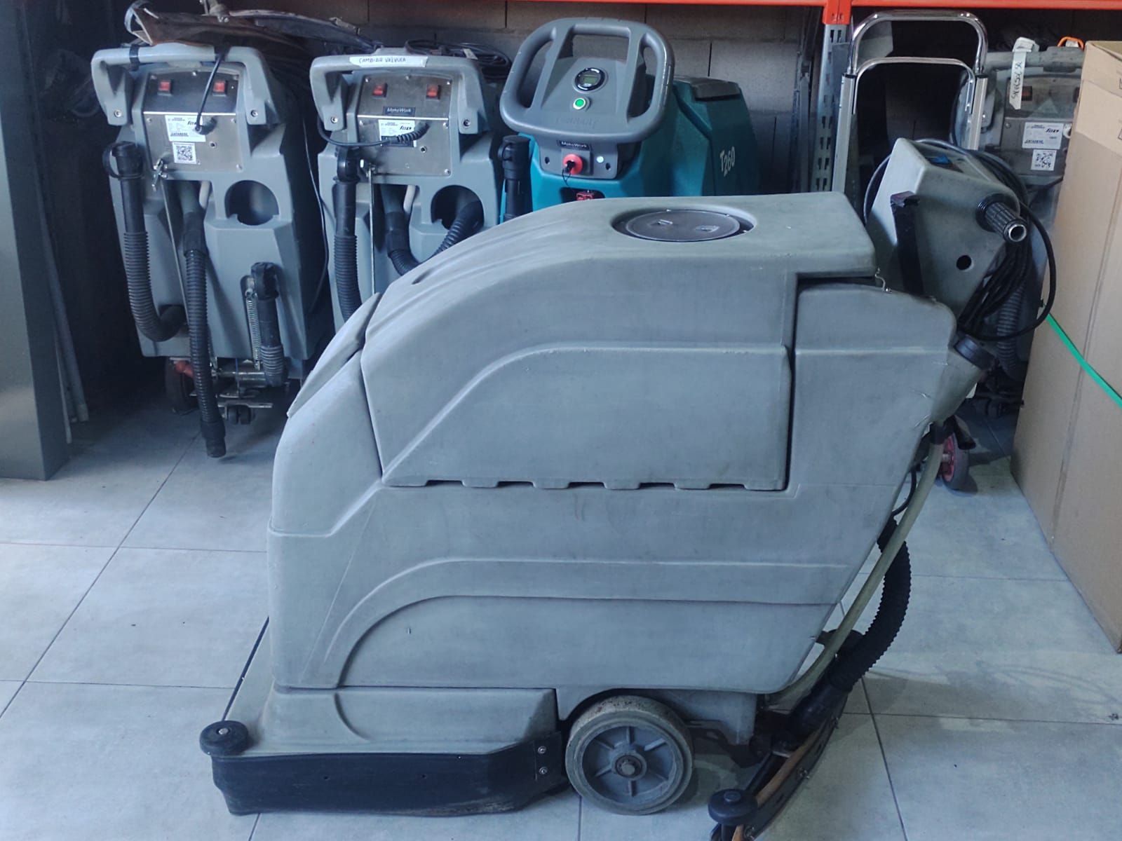 Máquina gris de limpieza de suelos en una zona de almacenamiento, con otros equipos de limpieza al fondo.