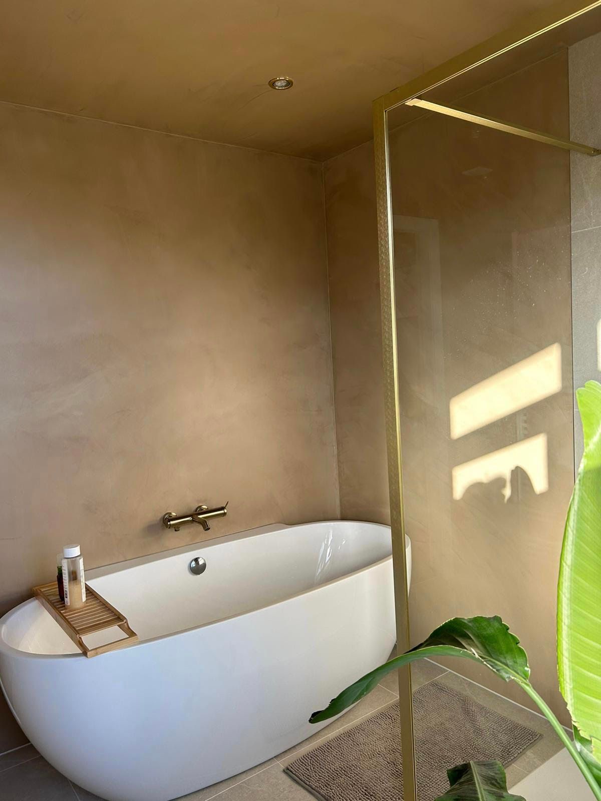 Moderne badkamer met vrijstaand wit bad, beige muren en een glazen douchewand in warm zonlicht.