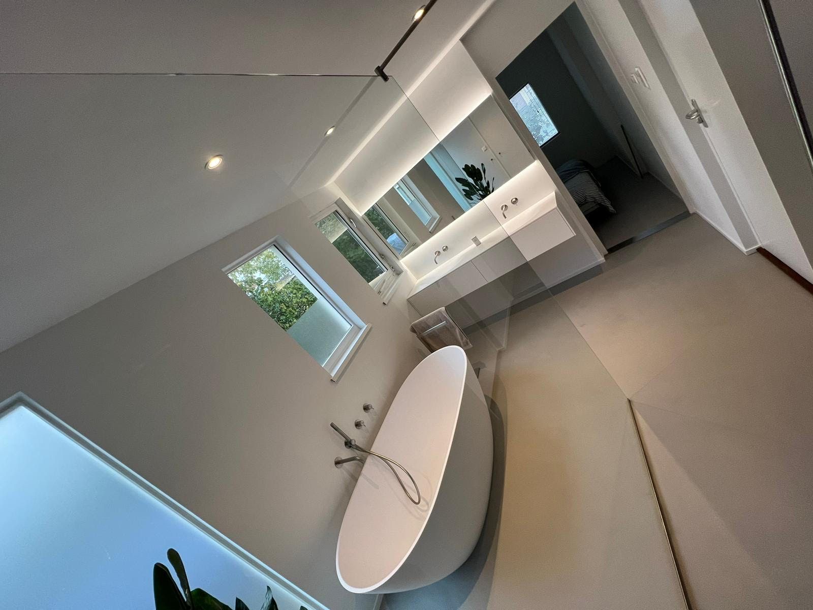 Moderne badkamer met een vrijstaand ovaal bad, groot raam, wastafelmeubel en neutrale grijze muren.