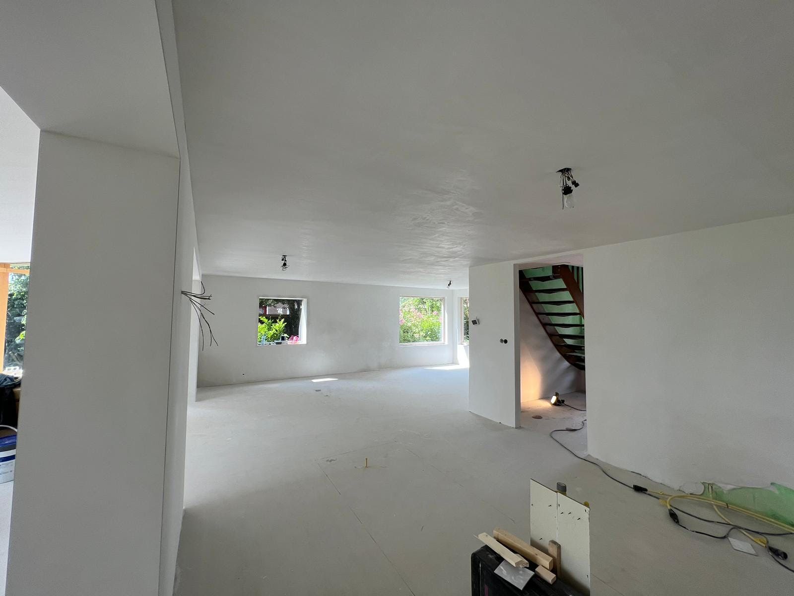 Een lege, witte binnenruimte in renovatie met een zichtbare trap, verspreid gereedschap en daglicht door de ramen.
