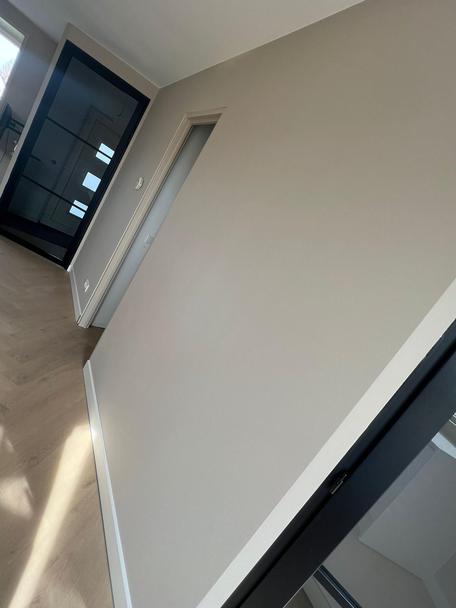 Lege beige kamer met een raam, radiator en donker deurkozijn, vanuit een lichte hoek bekeken.