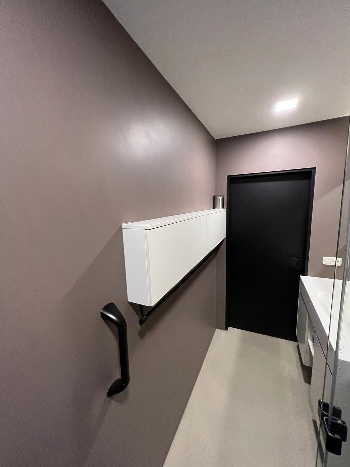Interieur van een toiletcabine met paarse muren, witte scheidingswanden, een wastafel en een gesloten zwarte deur.