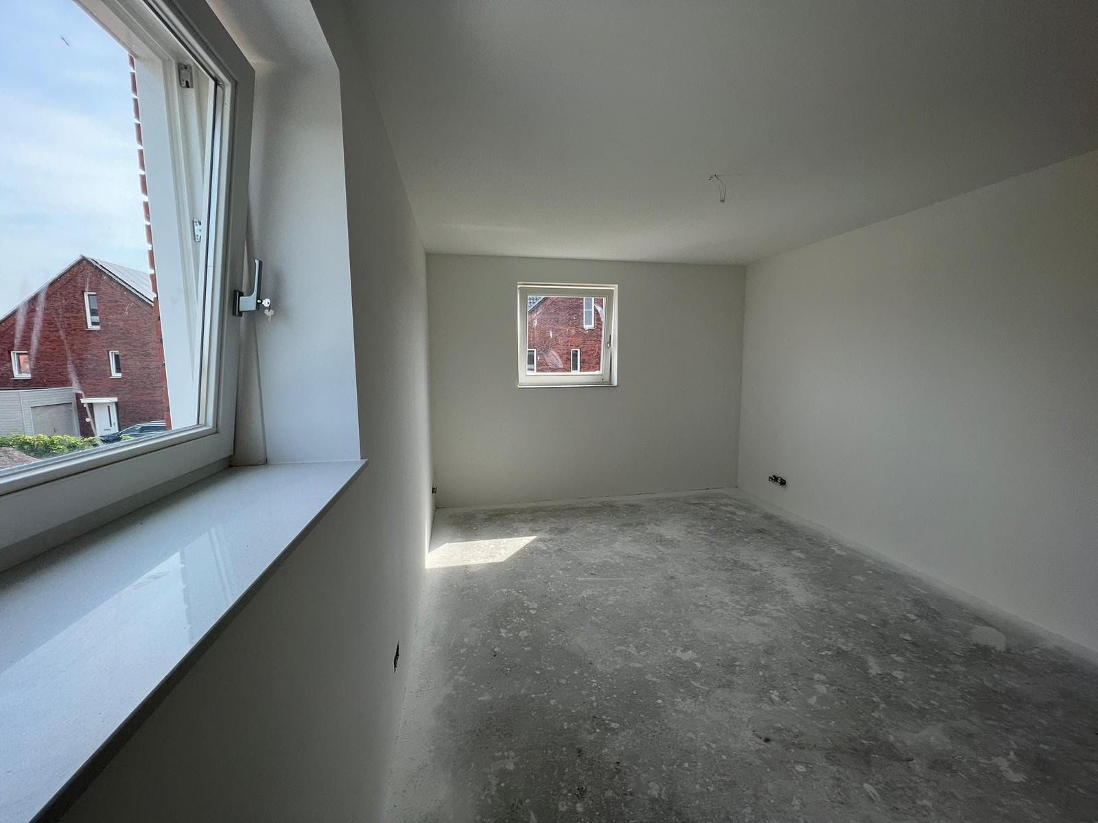 Een lege, onafgewerkte kamer met kale grijze muren, een betonnen vloer en ramen aan twee zijden.
