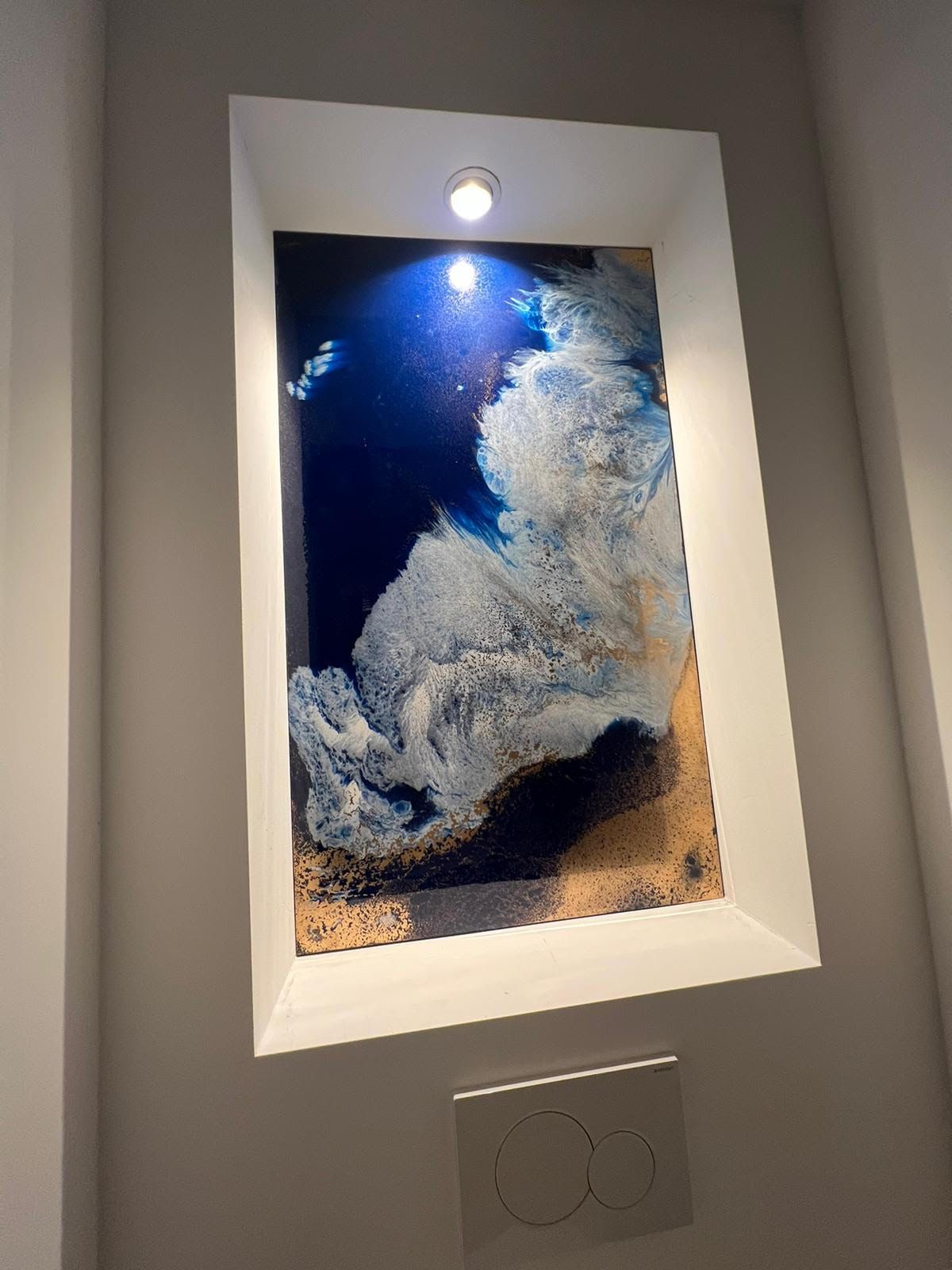 Ingelijst abstract blauw-wit kunstwerk, verlicht door een spot aan de muur boven een lichtschakelaar.