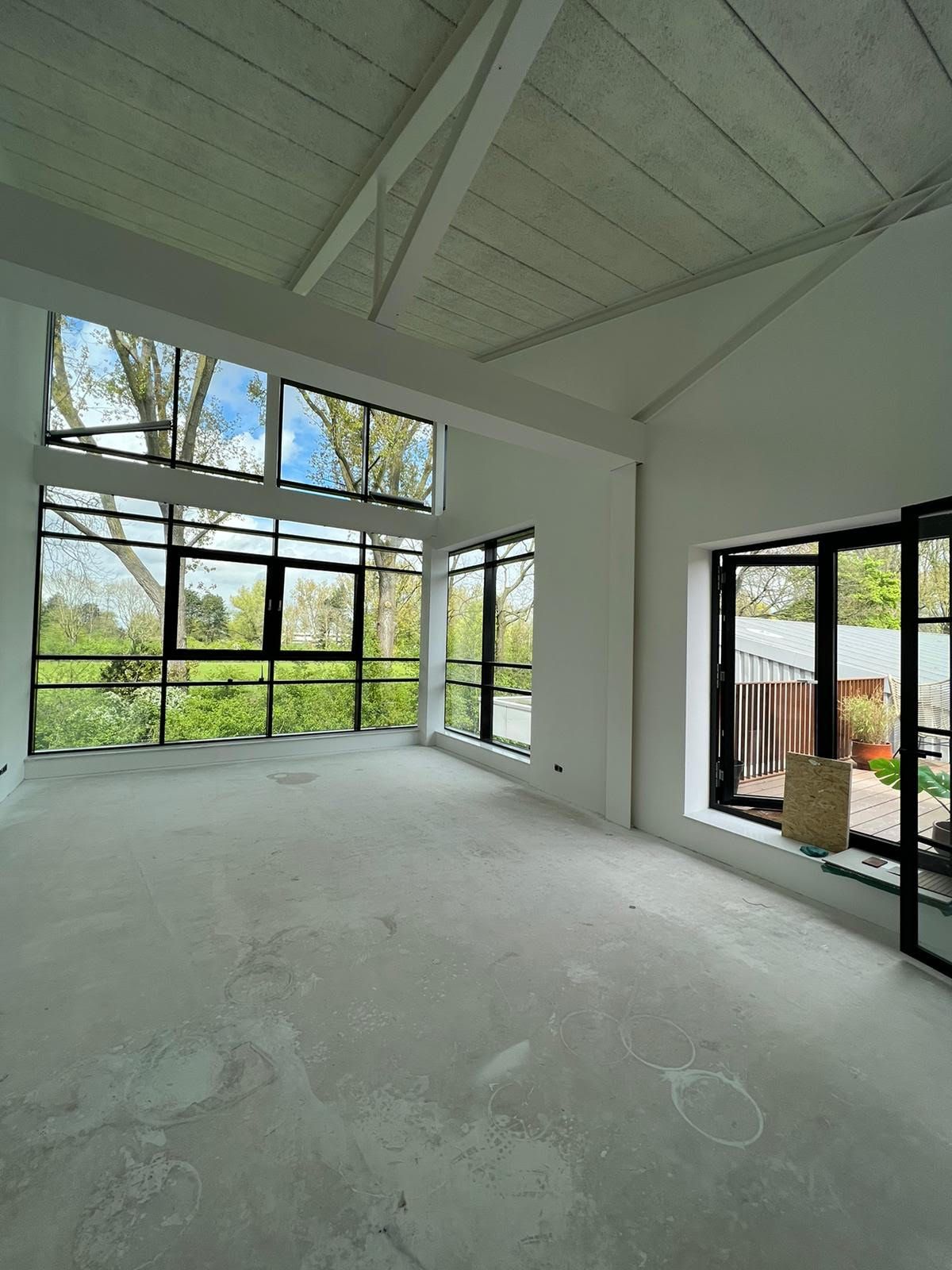 Lege, moderne kamer met hoog gewelfd plafond, grote ramen met zwarte kozijnen en een betonnen vloer.