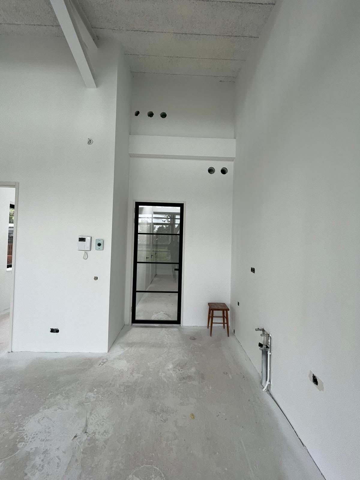 Een lege, witte kamer met een hoog plafond, een betonnen vloer en een glazen deur aan het uiteinde.