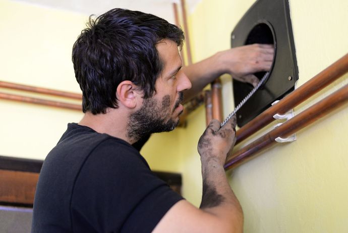 Un uomo, con il viso e le mani ricoperti di fuliggine, pulisce il camino, il muro giallo e i tubi di rame.
