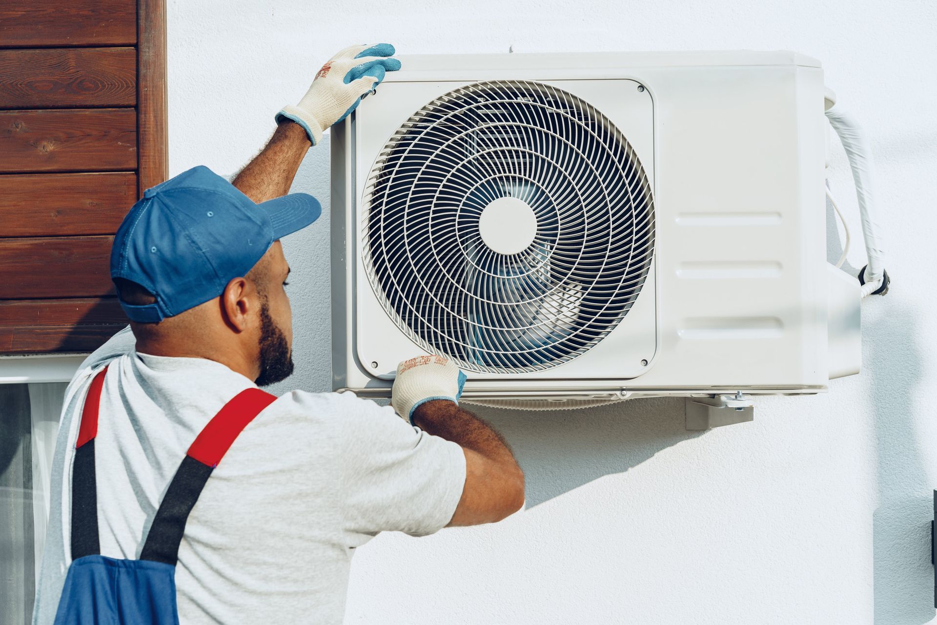 Technician servicing a wall-mounted outdoor air conditioner unit with tools and gloves