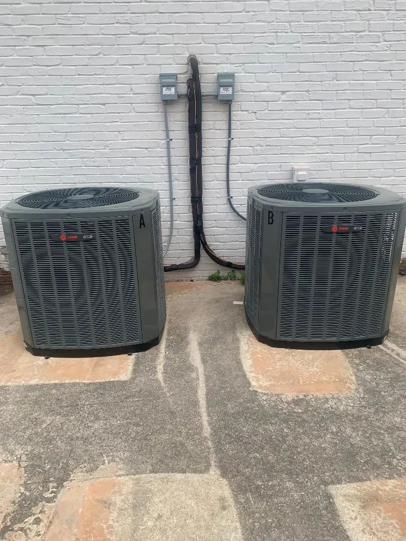 Two outdoor HVAC units beside a brick wall, with electrical conduit above them.