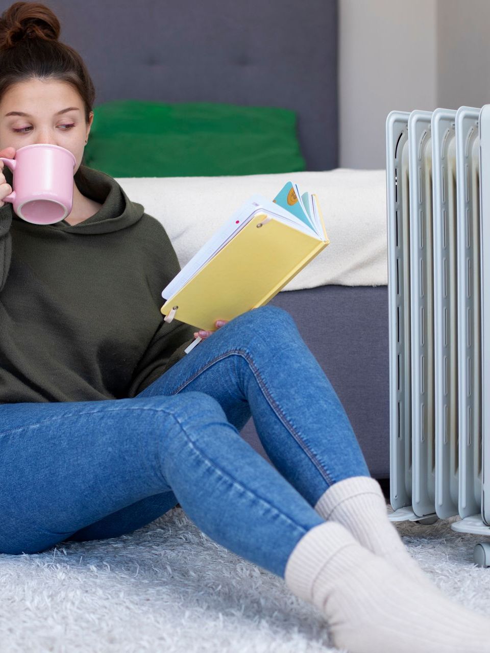 A woman is sitting on the floor reading a book and drinking coffee.