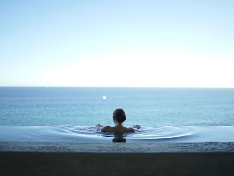 Pessoa relaxando em uma piscina infinita com vista para um oceano azul calmo sob um céu claro.