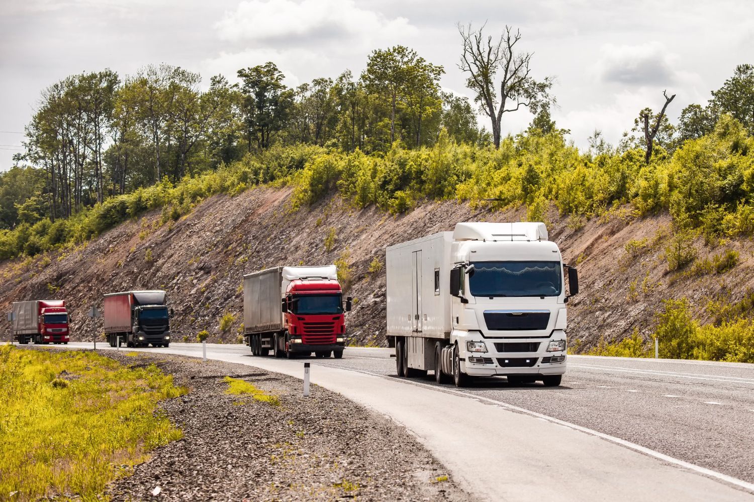 Truck convoy driving on a winding road beside a rocky hillside and trees