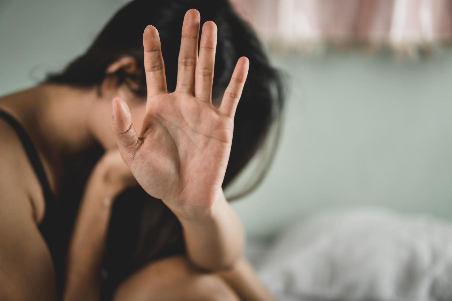 Person covering face with raised hand on a bed, with a blurred bedroom background