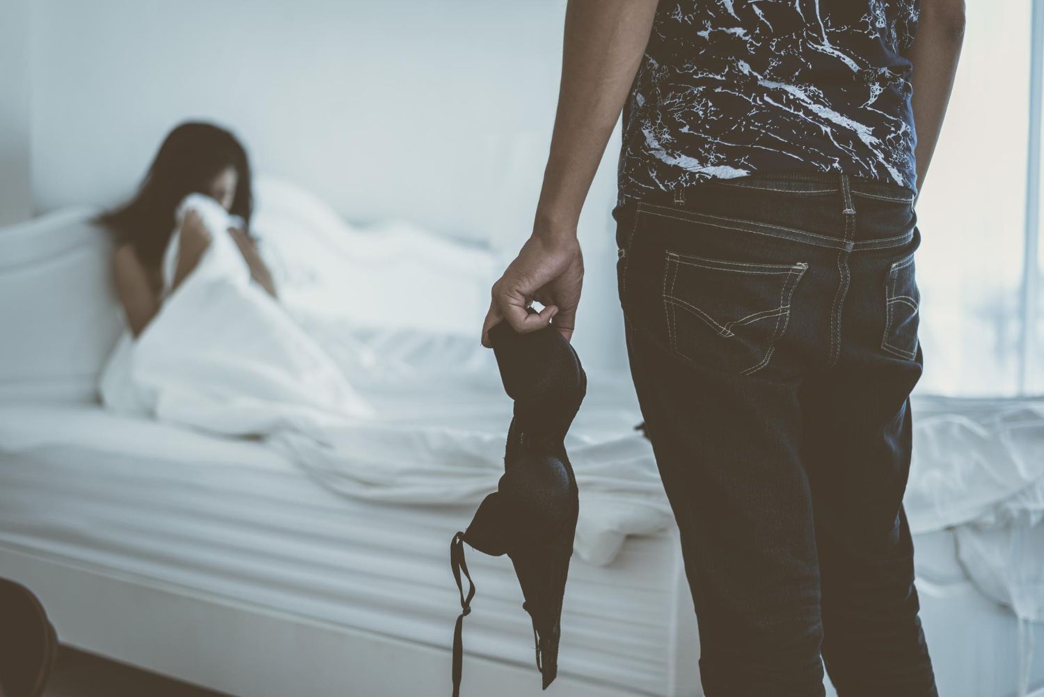 Man holding black lingerie in a bedroom, with a woman lying on a bed in the background