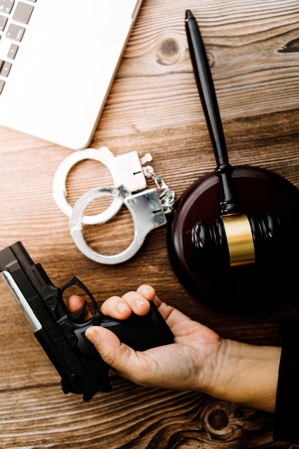 Hand holding a handgun beside handcuffs and a judge’s gavel on a wooden desk