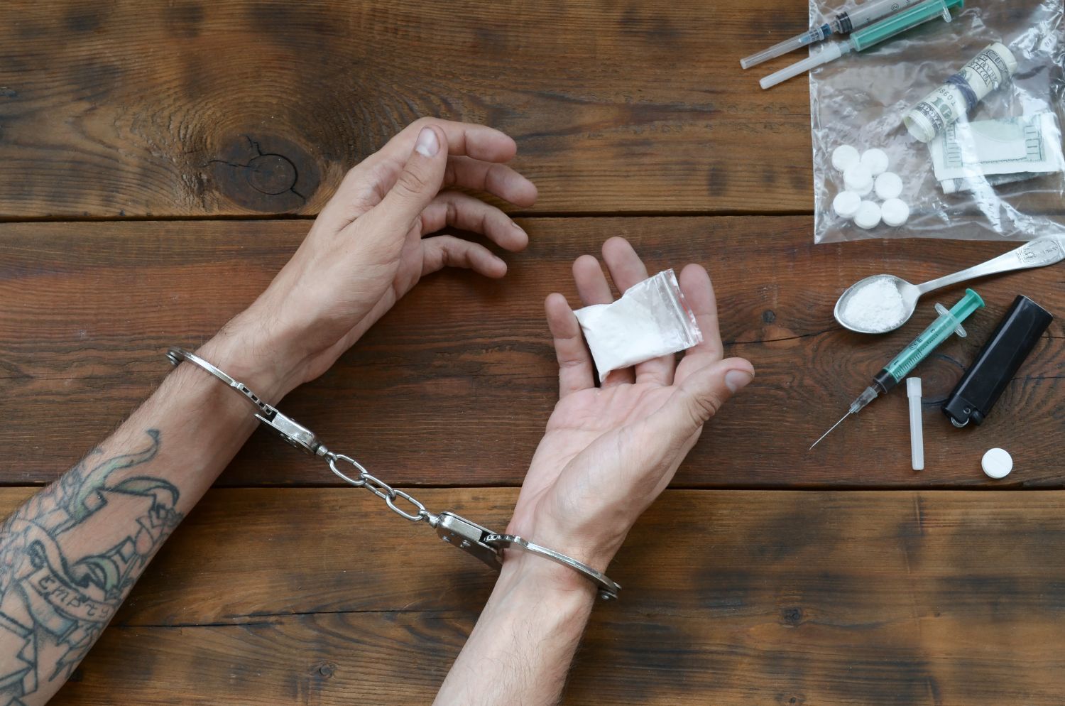 Handcuffed person holding a wrapped object on a wooden table with drug paraphernalia nearby