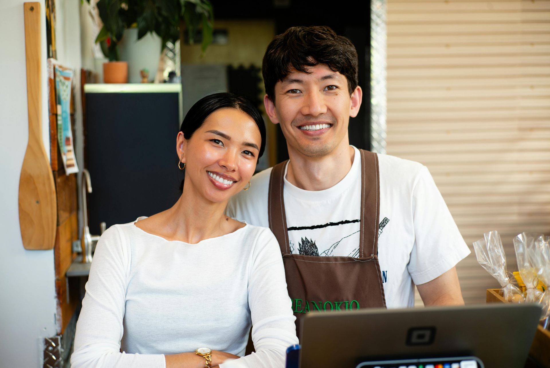 Two smiling people behind a counter in a café, with a laptop in front of them.