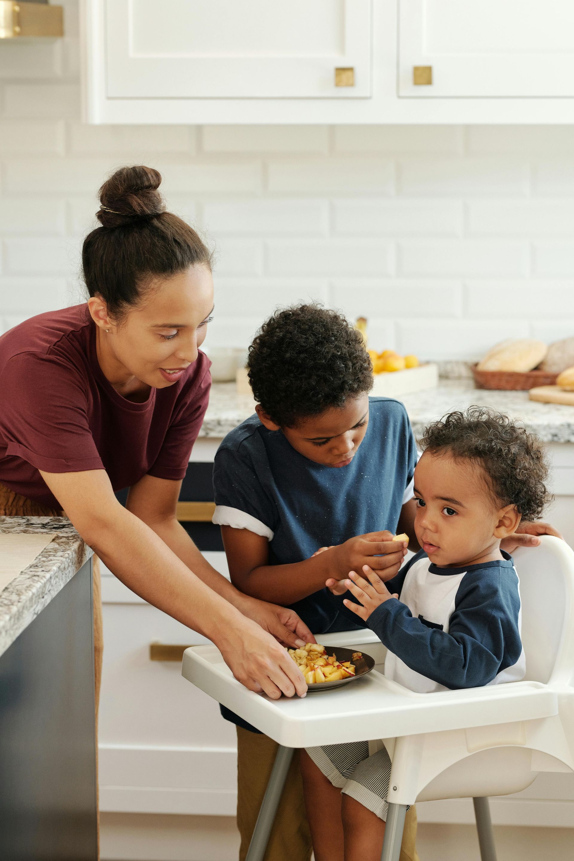 Parent feeds toddler at high chair while another child watches in a bright kitchen