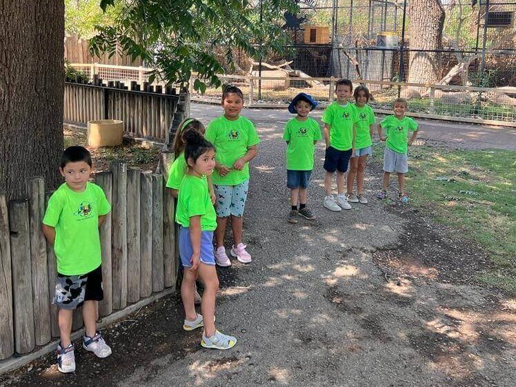 A group of children wearing green shirts are standing next to a wooden fence.