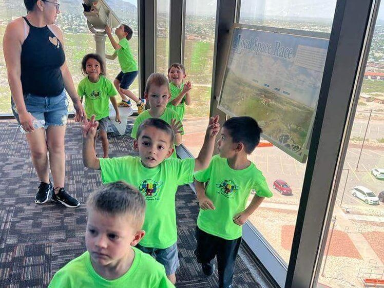 A group of children in green shirts are standing in front of a window.