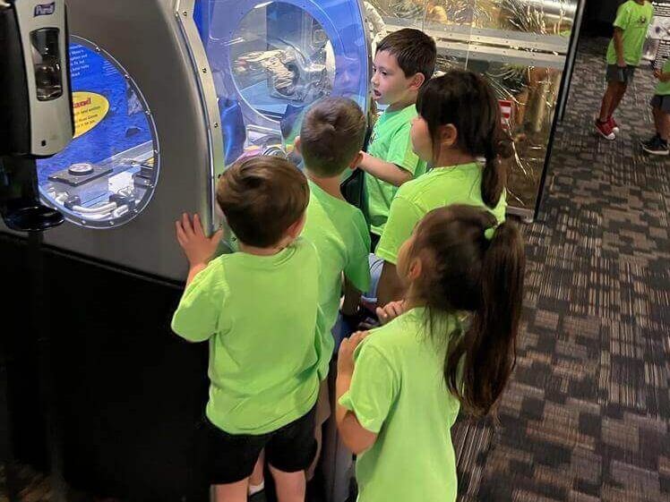A group of children in green shirts are looking at a machine.