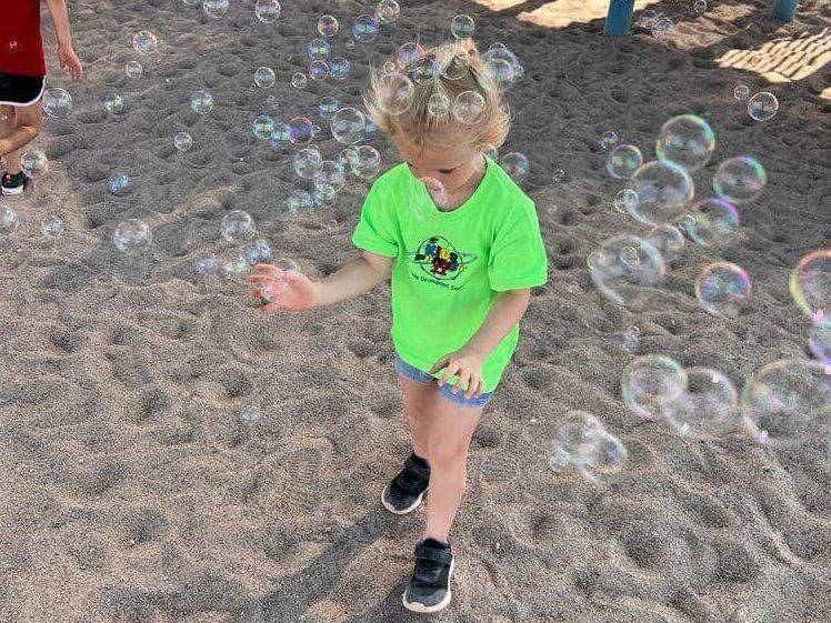 A little girl in a green shirt is playing with soap bubbles in the sand.