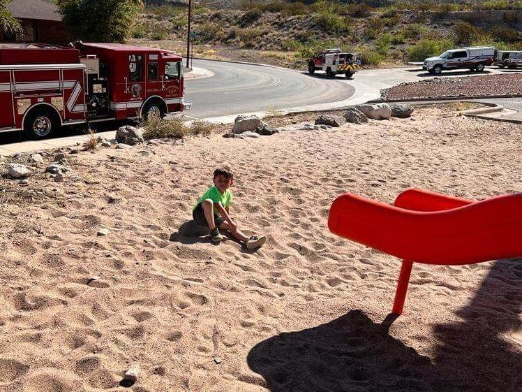 A little boy is playing in the sand with a fire truck in the background