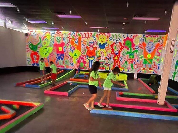 A group of children are playing mini golf in a room with neon painted walls.