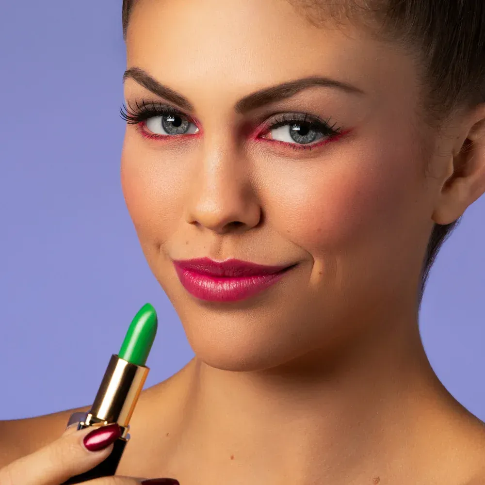 A person with red eyeliner and lipstick holds a tube of bright green lipstick against a solid purple background.