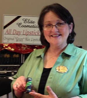 A woman in a green shirt smiles while holding a tube of Elite Cosmetics lipstick in front of a branded display sign.
