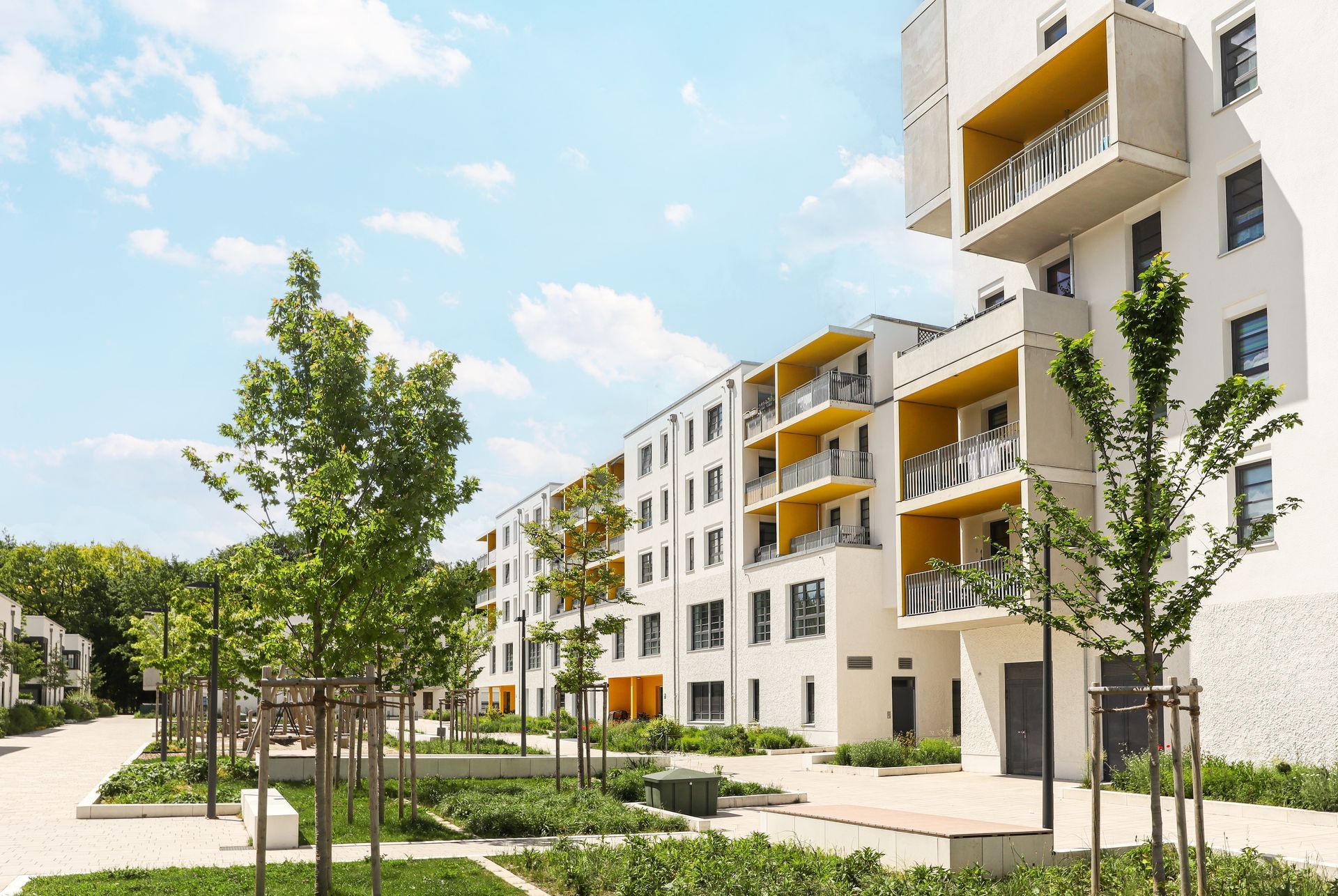 Modern white apartment complex with yellow accents, trees, and green landscaping under a bright blue sky.