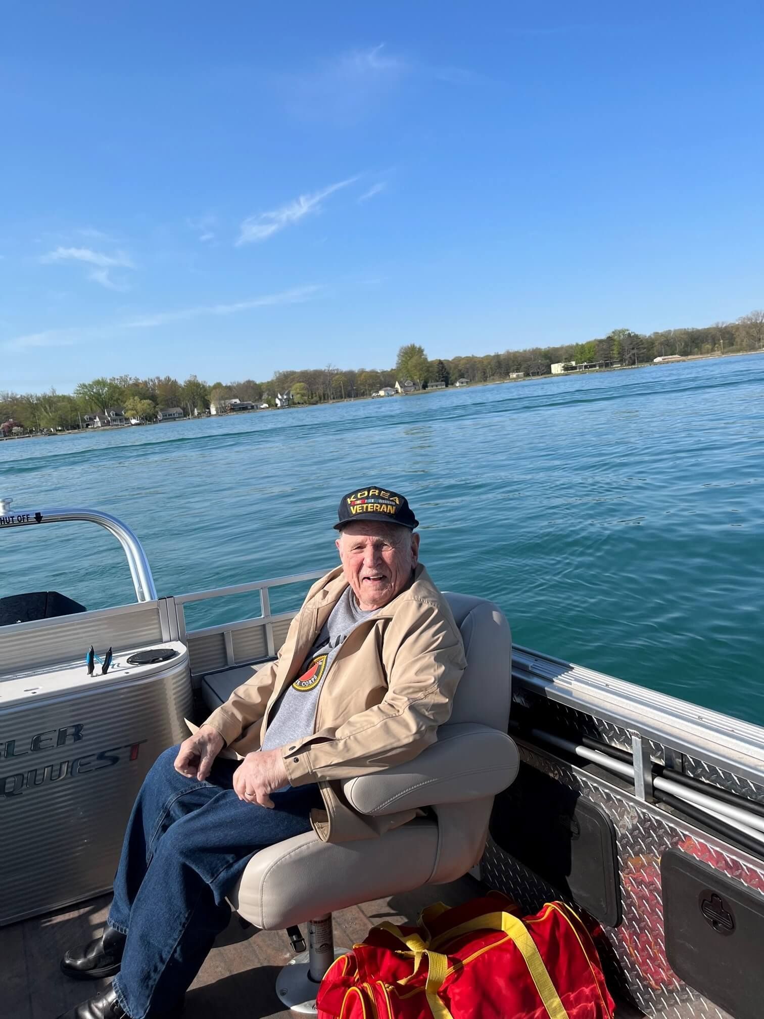 Man seated on a boat, on a body of water, wearing a cap and jacket, with blue skies in the background.