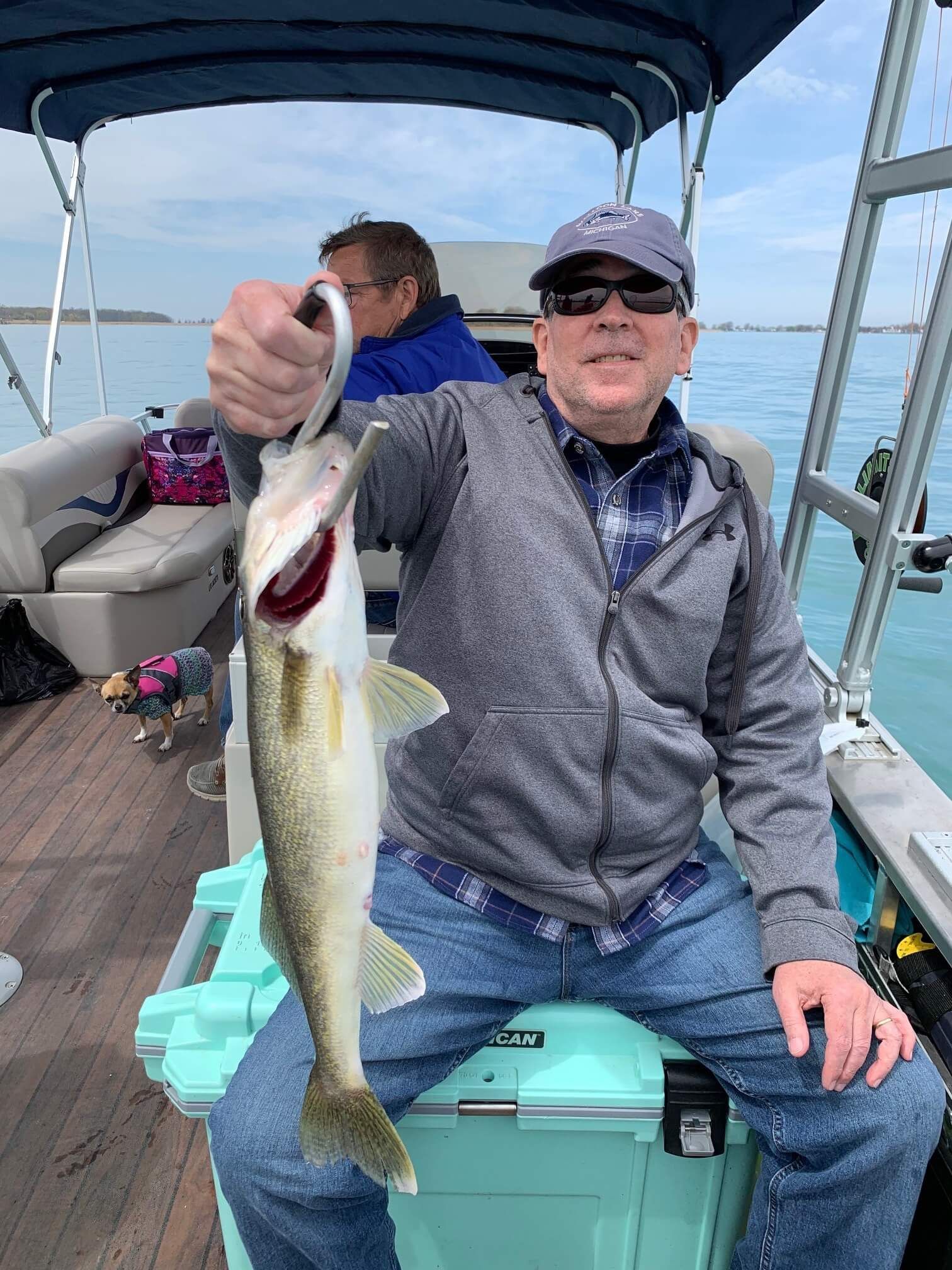 Man on a boat holds up a fish he caught. He smiles, a second person is behind him.