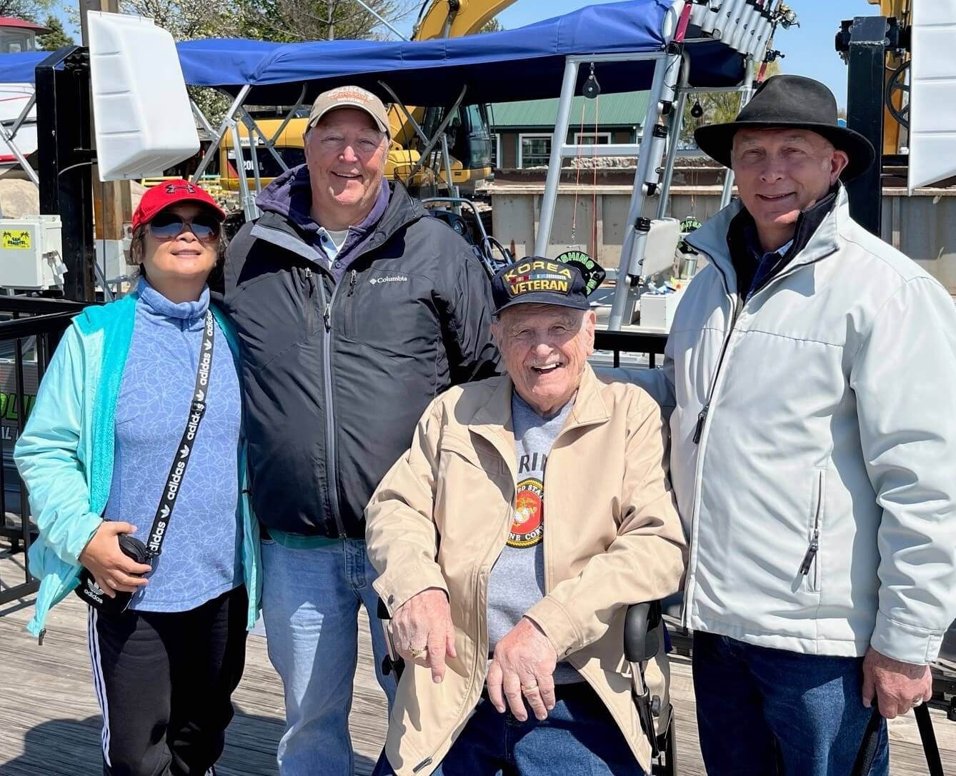 Four people posing outdoors, one in a wheelchair. Blue awning in background.