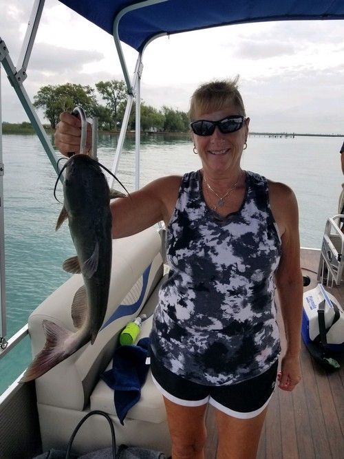 Woman on a boat holding up a catfish she caught. She wears sunglasses and a tank top. The background is water.