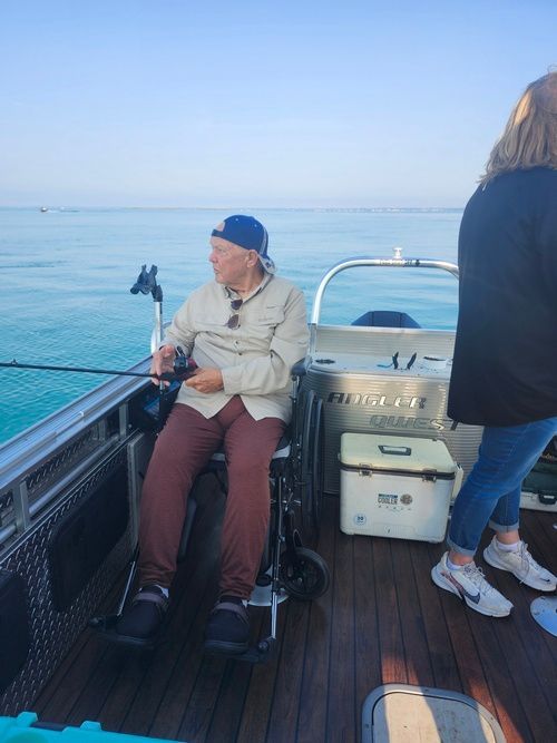Man in wheelchair fishing on a boat, looking at water. Another person standing nearby. Clear blue water and sky.