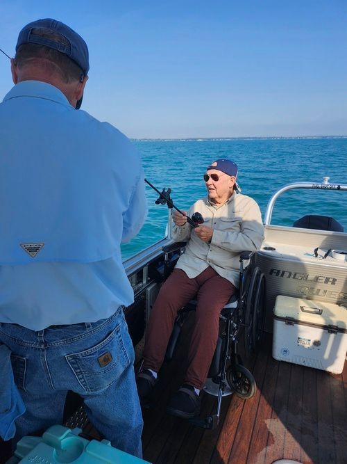 Man in wheelchair fishing on a boat with assistance; sunny day on the water.