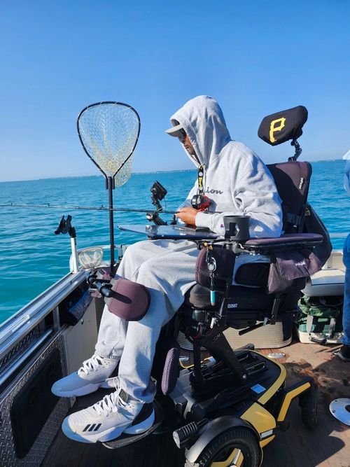 Person in wheelchair fishing on a boat on a sunny day, holding fishing rod, net nearby.
