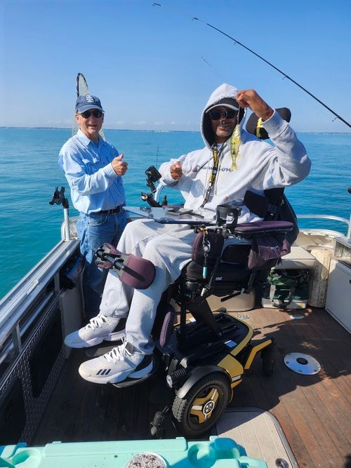 Man in wheelchair fishing on a boat, holding up a fish, with an older man beside him.
