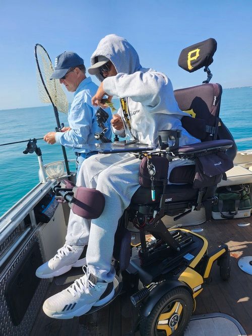 Man in wheelchair fishing on a boat; another man fishing beside him. Blue sky and water.