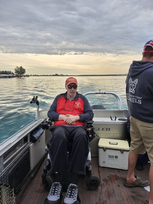 Man in wheelchair on a boat, wearing sunglasses and a red vest, on the water under a cloudy sky.
