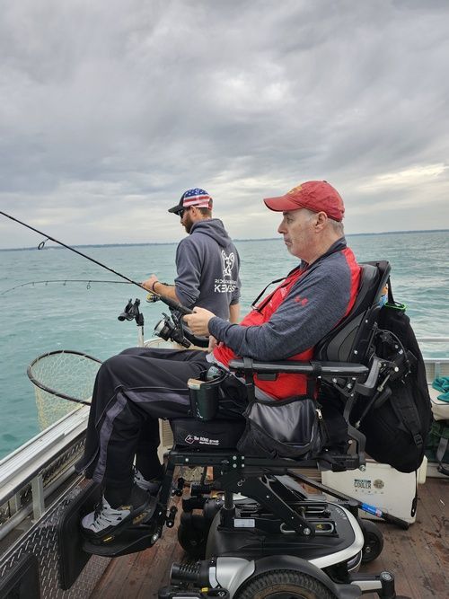 Man in wheelchair fishing on a boat with another person, cloudy sky.