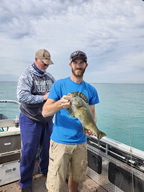 Man holding a fish on a boat; another man behind him. Clear water, cloudy sky.