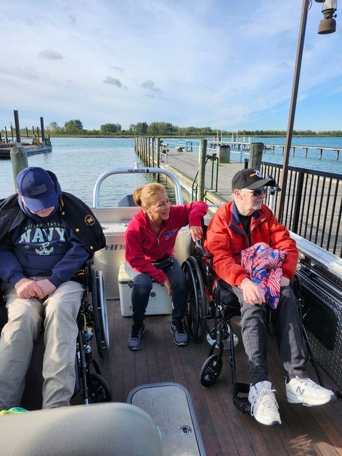Three people in wheelchairs on a pontoon boat near a pier; a woman leans towards one person. Sunny day.
