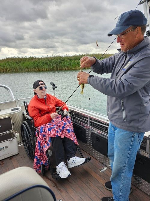 Person in wheelchair fishing on a boat, assisted by another person holding a fish on a fishing rod. Outdoors.