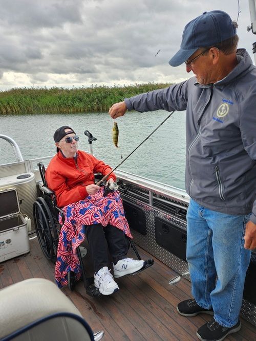 Man on a pontoon boat fishing, seated person in wheelchair looks on. The other man is holding up small fish.