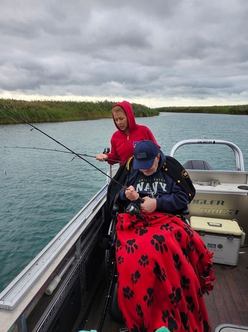 Person in wheelchair fishing on a boat with assistance. Overcast day.