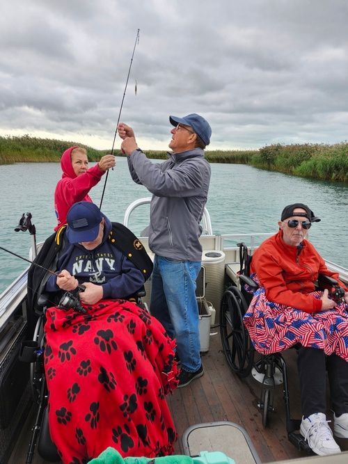 People in wheelchairs fishing from a boat on a lake under an overcast sky.
